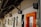 A corridor with a wooden ceiling and multiple signs on the wall, including a medical cross, a nursery room symbol, and other signs indicating various rooms and services. There is an orange-framed door on the right side and several windows along the wall.