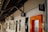 A corridor with a wooden ceiling and multiple signs on the wall, including a medical cross, a nursery room symbol, and other signs indicating various rooms and services. There is an orange-framed door on the right side and several windows along the wall.