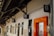 A corridor with a wooden ceiling and multiple signs on the wall, including a medical cross, a nursery room symbol, and other signs indicating various rooms and services. There is an orange-framed door on the right side and several windows along the wall.
