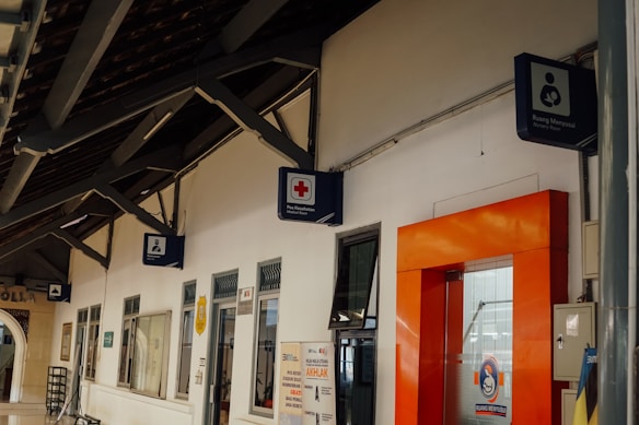 A corridor with a wooden ceiling and multiple signs on the wall, including a medical cross, a nursery room symbol, and other signs indicating various rooms and services. There is an orange-framed door on the right side and several windows along the wall.