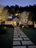 A charming house set against a backdrop of tall trees, with warm lighting illuminating its exterior. A stone pathway leads up to the house, framed by neatly trimmed bushes and trees. The overall scene is serene and inviting.