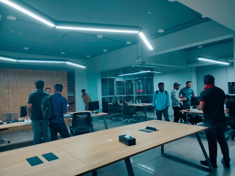 A modern office space with several people gathered around desks. The room is well-lit with fluorescent lights, featuring wooden tables and computer screens. Some individuals are standing and conversing while others are seated. Glass walls separate part of the space, adding to the open, collaborative atmosphere.