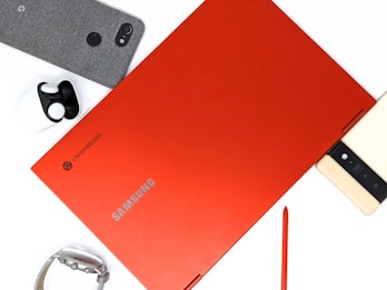 A red Samsung Chromebook laptop is positioned centrally with its brand name visible. Surrounding it are various tech accessories, including a smartphone with a fabric cover, a pair of wireless earbuds, a smart watch, and a stylus pen.