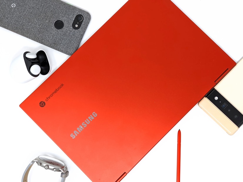 A red Samsung Chromebook laptop is positioned centrally with its brand name visible. Surrounding it are various tech accessories, including a smartphone with a fabric cover, a pair of wireless earbuds, a smart watch, and a stylus pen.