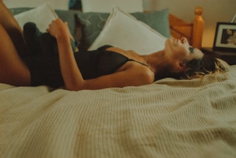 A person is lying on a bed, wearing black nightwear. The setting appears to be a bedroom with a soft, intimate atmosphere. There is a picture frame visible in the background, and the bed is covered with a beige striped blanket. The lighting is warm and subdued, contributing to a calm and reflective mood.