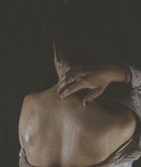 A person is facing away, revealing their bare back with water droplets on their skin. One hand rests gently on their shoulder, with a ring visible on the finger. The lighting is dim, creating a moody and intimate atmosphere.