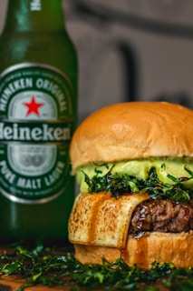 A close-up of a gourmet hamburger with a thick beef patty, melted cheese, and fresh greens, paired with a bottle of Heineken beer in the background. The burger showcases a glossy bun and juicy filling, indicating a rich and indulgent meal.