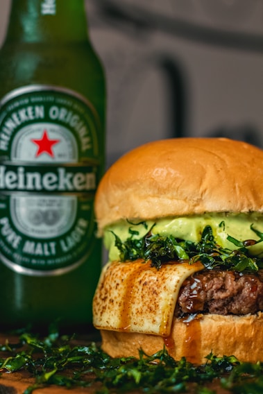 A close-up of a gourmet hamburger with a thick beef patty, melted cheese, and fresh greens, paired with a bottle of Heineken beer in the background. The burger showcases a glossy bun and juicy filling, indicating a rich and indulgent meal.