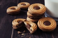 A stack of round cookies with a central chocolate filling is placed next to a glass of milk on a dark, wooden surface. Some cookies are scattered, and crumbs are visibly present, suggesting a freshly served snack.