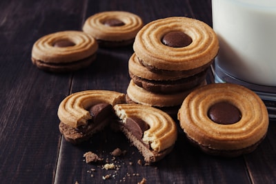 A stack of round cookies with a central chocolate filling is placed next to a glass of milk on a dark, wooden surface. Some cookies are scattered, and crumbs are visibly present, suggesting a freshly served snack.