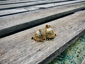 A pair of intricately designed, gold-colored earrings with dome shapes and embellishments resting on a wooden surface. The wood has a weathered appearance with visible grains and lines running through it.