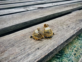 A pair of intricately designed, gold-colored earrings with dome shapes and embellishments resting on a wooden surface. The wood has a weathered appearance with visible grains and lines running through it.