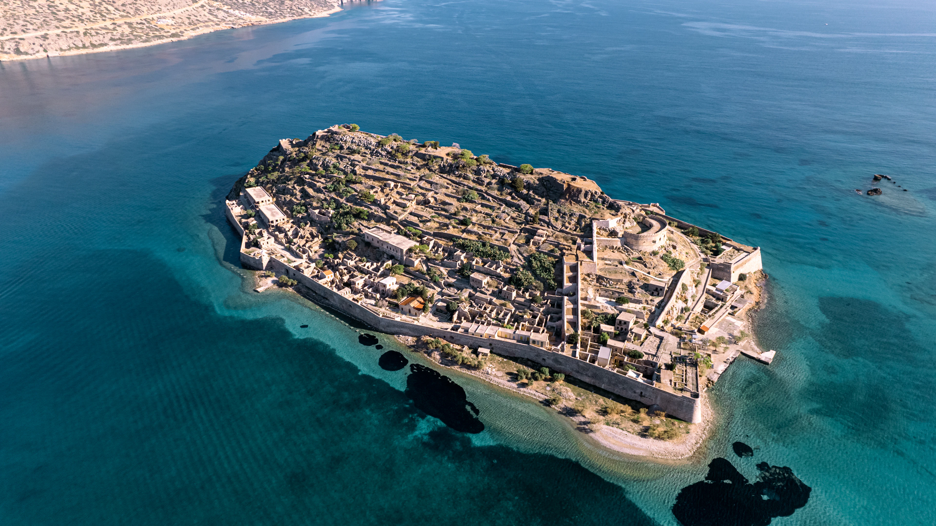 The fortified island of Spinalonga seen from the air, its Venetian bastion walls and the roofless lanes of the abandoned leper colony ringed by clear turquoise shallows off the Cretan coast.