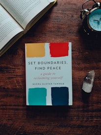 A wooden table holds an open book, an alarm clock, a crystal, and a self-help book titled 'Set Boundaries, Find Peace'. The book cover features colorful swatches in yellow, red, teal, and blue.