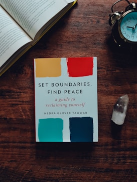 A wooden table holds an open book, an alarm clock, a crystal, and a self-help book titled 'Set Boundaries, Find Peace'. The book cover features colorful swatches in yellow, red, teal, and blue.