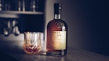 A bottle of Monkey Shoulder blended malt Scotch whisky is placed on a wooden table next to a crystal glass filled with the same whisky. The background includes a blurred section of a shelf with glasses, creating an elegant and cozy atmosphere.