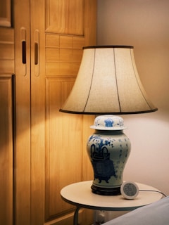 A ceramic table lamp with a beige lampshade sits on a small round table. The lamp has a blue and white pattern, resembling traditional Chinese designs. A digital clock displaying temperature and humidity is placed beside the lamp. Wooden closet doors form the background.