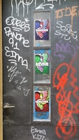 A dark door with various graffiti and colorful posters. The graffiti includes words and abstract symbols in white, red, and orange. Three vibrant posters with the words 'Reimagine,' 'Revive,' and 'Rediscover' feature colorful, artistic designs using red, green, and purple hues, each with abstract shapes and patterns.