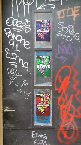 A dark door with various graffiti and colorful posters. The graffiti includes words and abstract symbols in white, red, and orange. Three vibrant posters with the words 'Reimagine,' 'Revive,' and 'Rediscover' feature colorful, artistic designs using red, green, and purple hues, each with abstract shapes and patterns.