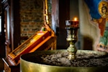A dimly lit church interior features a brass candle holder in the foreground, topped with a lit candle. The background displays an icon painting with vibrant colors and an exposed brick wall. The atmosphere is serene and reverential.
