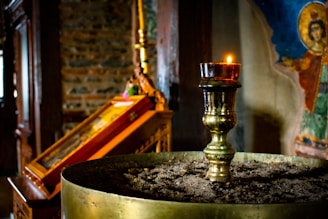 A dimly lit church interior features a brass candle holder in the foreground, topped with a lit candle. The background displays an icon painting with vibrant colors and an exposed brick wall. The atmosphere is serene and reverential.
