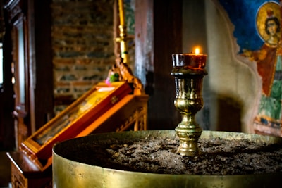 A dimly lit church interior features a brass candle holder in the foreground, topped with a lit candle. The background displays an icon painting with vibrant colors and an exposed brick wall. The atmosphere is serene and reverential.