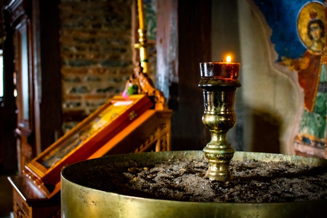 A dimly lit church interior features a brass candle holder in the foreground, topped with a lit candle. The background displays an icon painting with vibrant colors and an exposed brick wall. The atmosphere is serene and reverential.