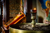 A dimly lit church interior features a brass candle holder in the foreground, topped with a lit candle. The background displays an icon painting with vibrant colors and an exposed brick wall. The atmosphere is serene and reverential.