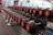 A row of neatly organized dumbbells with visible weight labels on a rack, set in a gym environment. The flooring and walls feature a mix of colors, and exercise machines are visible in the background.