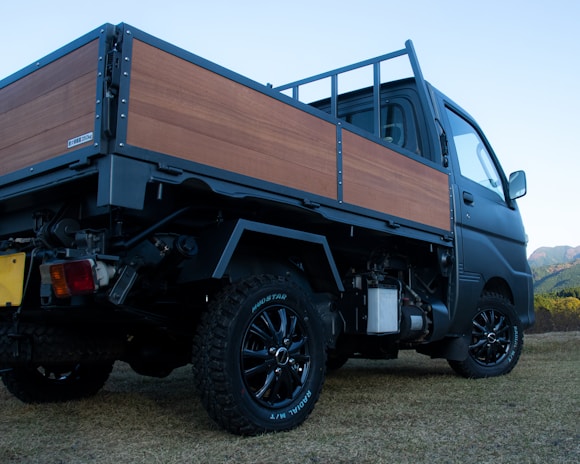 A small pickup truck with a flatbed covered with wooden panels is parked on a grassy area. The truck has black wheels with rugged tires and is seen from a low angle. In the background, there are hills and a clear sky indicating an outdoor and possibly rural setting.