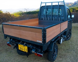 A small truck with a wooden flatbed parked on grassy terrain. The vehicle has black wheels and a distinct license plate. The background features lush green hills and distant mountains under a clear sky.