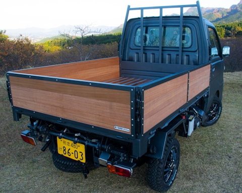 A small truck with a wooden flatbed parked on grassy terrain. The vehicle has black wheels and a distinct license plate. The background features lush green hills and distant mountains under a clear sky.