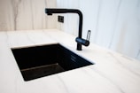 A modern kitchen sink setup with a matte black faucet and a matching black rectangular sink set in a white marble countertop. The faucet has a minimalist design with a sleek handle. The surrounding area shows clean, white, marble-like surface creating a contemporary and elegant look.