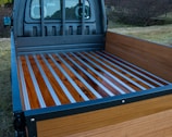 The image shows the bed of a truck with a wooden floor and metal strips running lengthwise. The truck bed is surrounded by wooden sides and set against an outdoor background with grass and trees.