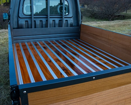 The image shows the bed of a truck with a wooden floor and metal strips running lengthwise. The truck bed is surrounded by wooden sides and set against an outdoor background with grass and trees.