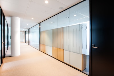 A modern office corridor with glass walls dividing individual rooms. The corridor features a clean design with white ceilings and walls, beige carpeted floors, and a prominent white column. Ceiling lights provide bright illumination, and there is a minimalistic decor with an emphasis on spaciousness and transparency.