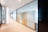 A modern office corridor with glass walls dividing individual rooms. The corridor features a clean design with white ceilings and walls, beige carpeted floors, and a prominent white column. Ceiling lights provide bright illumination, and there is a minimalistic decor with an emphasis on spaciousness and transparency.