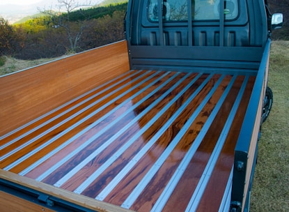 A truck bed with a sleek design features polished wooden planks framed by metal strips. The truck is parked in a grassy area with a scenic view of trees and hills in the background.