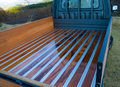 A truck bed with a sleek design features polished wooden planks framed by metal strips. The truck is parked in a grassy area with a scenic view of trees and hills in the background.