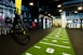 A modern gym space featuring a green track laid out on the floor with numbered lanes. TRX suspension trainers hang prominently in the foreground. The gym is equipped with various exercise equipment, including kettlebells and punching bags. In the background, there is a FIT sign near a reception desk under a low-ceilinged industrial design.