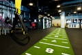 A modern gym space featuring a green track laid out on the floor with numbered lanes. TRX suspension trainers hang prominently in the foreground. The gym is equipped with various exercise equipment, including kettlebells and punching bags. In the background, there is a FIT sign near a reception desk under a low-ceilinged industrial design.