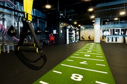 A modern gym space featuring a green track laid out on the floor with numbered lanes. TRX suspension trainers hang prominently in the foreground. The gym is equipped with various exercise equipment, including kettlebells and punching bags. In the background, there is a FIT sign near a reception desk under a low-ceilinged industrial design.