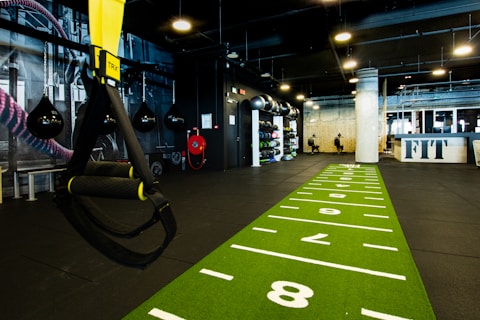 A modern gym space featuring a green track laid out on the floor with numbered lanes. TRX suspension trainers hang prominently in the foreground. The gym is equipped with various exercise equipment, including kettlebells and punching bags. In the background, there is a FIT sign near a reception desk under a low-ceilinged industrial design.