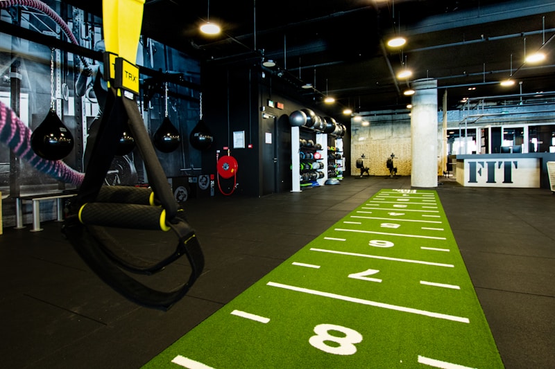 A modern gym space featuring a green track laid out on the floor with numbered lanes. TRX suspension trainers hang prominently in the foreground. The gym is equipped with various exercise equipment, including kettlebells and punching bags. In the background, there is a FIT sign near a reception desk under a low-ceilinged industrial design.