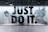 A large, bold slogan 'JUST DO IT.' is displayed with an action-packed background featuring a blurred image of a person running. The wall is part of an interior with a modern design, and there is a sign on the right side welcoming people to Nike, highlighting various aspects like experts, sport, and rewards. The color scheme is predominantly monochrome.