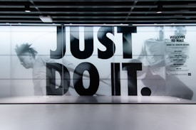 A large, bold slogan 'JUST DO IT.' is displayed with an action-packed background featuring a blurred image of a person running. The wall is part of an interior with a modern design, and there is a sign on the right side welcoming people to Nike, highlighting various aspects like experts, sport, and rewards. The color scheme is predominantly monochrome.
