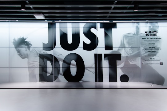 A large, bold slogan 'JUST DO IT.' is displayed with an action-packed background featuring a blurred image of a person running. The wall is part of an interior with a modern design, and there is a sign on the right side welcoming people to Nike, highlighting various aspects like experts, sport, and rewards. The color scheme is predominantly monochrome.