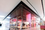 A modern retail store with a sleek glass facade located inside a shopping mall. The store features a vibrant interior with prominent red and pink lighting, creating a contemporary and dynamic appeal. The entrance is spacious and welcoming, with clear visibility of products displayed inside.