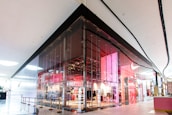 A modern retail store with a sleek glass facade located inside a shopping mall. The store features a vibrant interior with prominent red and pink lighting, creating a contemporary and dynamic appeal. The entrance is spacious and welcoming, with clear visibility of products displayed inside.