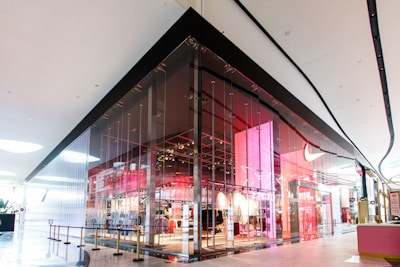 A modern retail store with a sleek glass facade located inside a shopping mall. The store features a vibrant interior with prominent red and pink lighting, creating a contemporary and dynamic appeal. The entrance is spacious and welcoming, with clear visibility of products displayed inside.